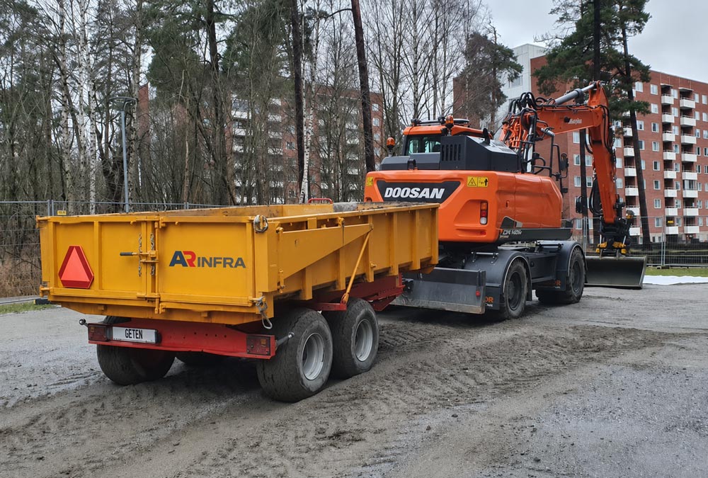 En orange grävmaskin är parkerad bredvid en gul släpvagn på en grusig byggarbetsplats